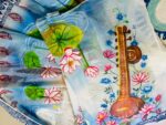 Saraswati theme hand painted artificial muslin saree from KhatiBaazaar featuring a traditional veena and pink lotus design on a white and blue gradient backdrop