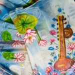 Saraswati theme hand painted artificial muslin saree from KhatiBaazaar featuring a traditional veena and pink lotus design on a white and blue gradient backdrop