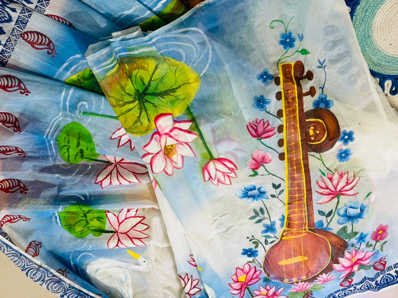 saraswati-theme-hand-painted-muslin-saree-khatibaazaar Saraswati theme hand painted artificial muslin saree from KhatiBaazaar featuring a traditional veena and pink lotus design on a white and blue gradient backdrop